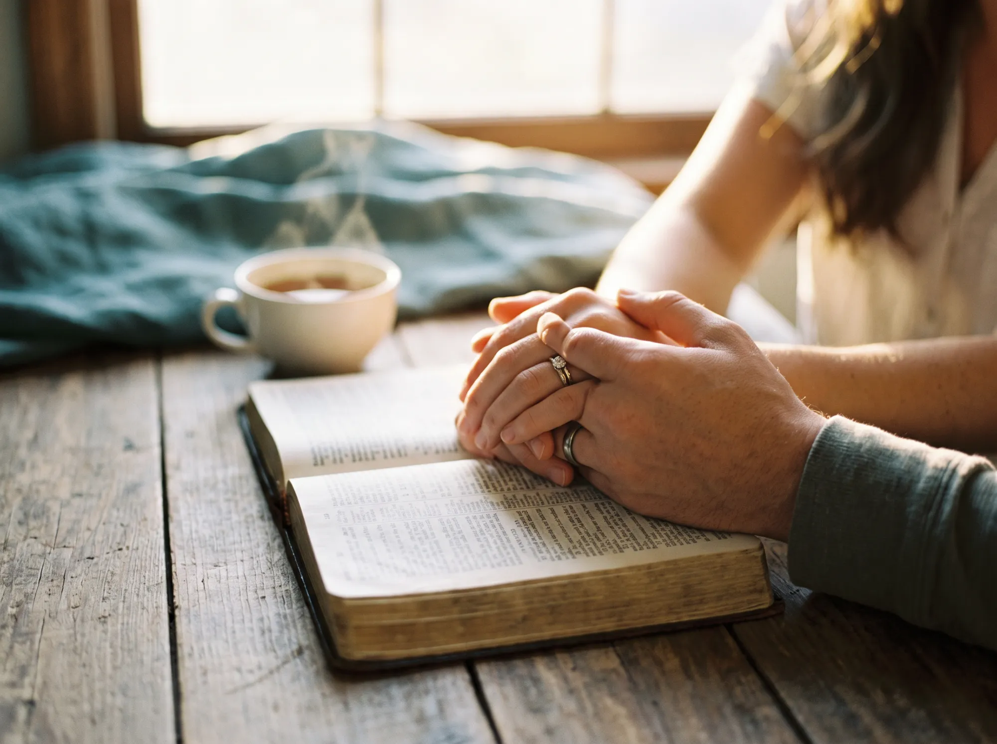 Clasped hands with wedding rings resting on an open Bible beside a cup of tea
